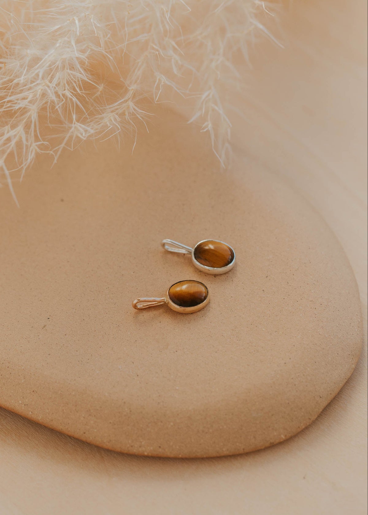 Pair of gold pendants on a beige stone surface with white dried leaves in the background
