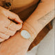Close-up of hands wearing gold rings and a watch, with a brown garment in the background.
