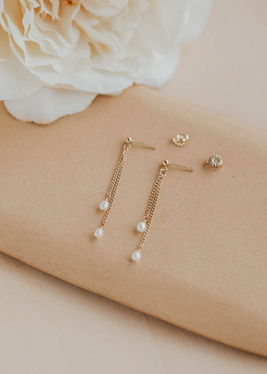 Gold earrings with pearl drops on a beige surface with a white flower.