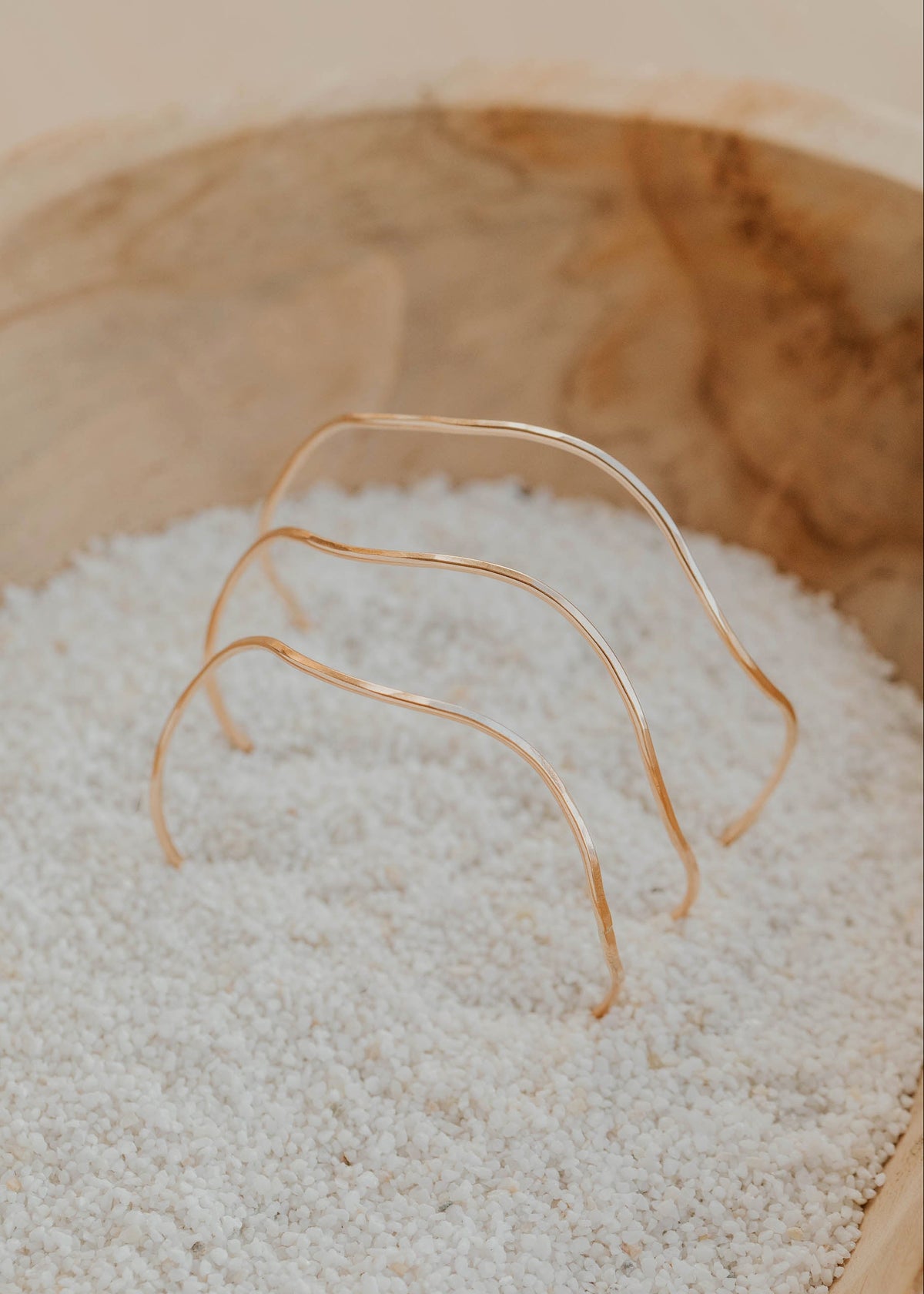 Three gold bangle bracelets on a bed of white sand inside a wooden bowl.