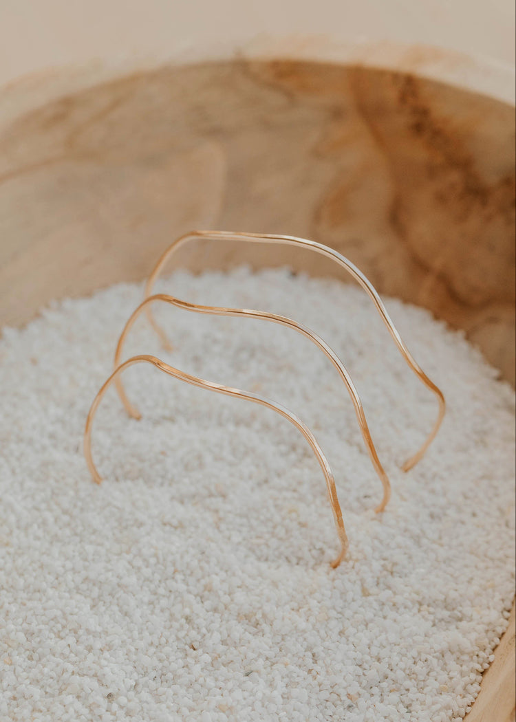 Three gold bangle bracelets on a bed of white sand inside a wooden bowl.