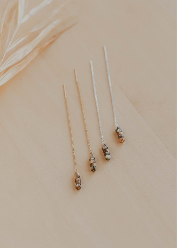 Three delicate earrings on a beige surface with a blurred leaf in the background